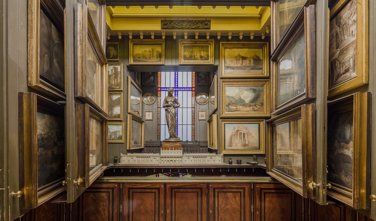 Picture Room Recess at Sir John Soane Museum. Art-filled room with framed paintings, statue, and diorama under a stained glass window.