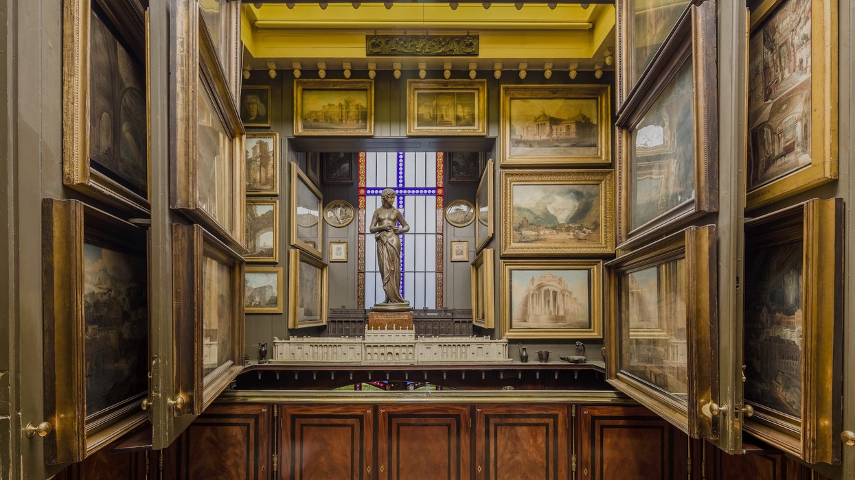 Picture Room Recess at Sir John Soane Museum. Art-filled room with framed paintings, statue, and diorama under a stained glass window.