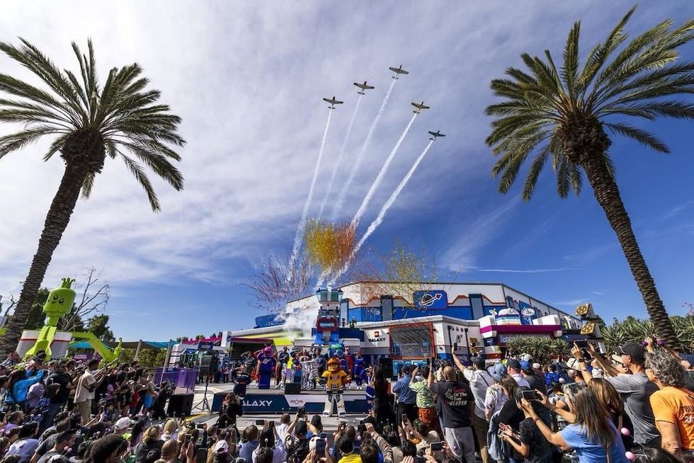 Planes fly overhead, colorful smoke and confetti burst, crowd gathers under palm trees.