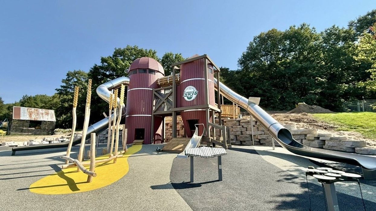 Playground with slides and climbing structures, surrounded by trees and a sunny sky.