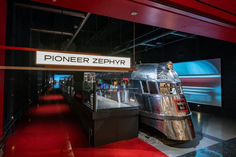 Red and black hallway with lit sign that reads "pioneer zephyr" and chrome passenger train at right