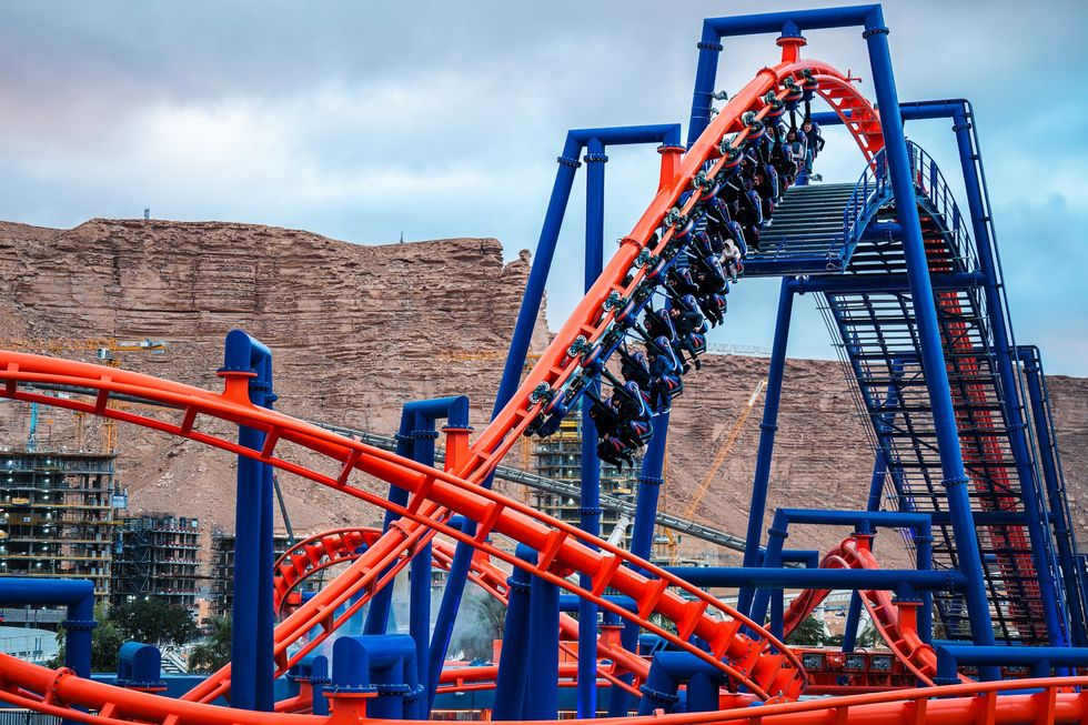 Red and blue roller coaster with riders, set against rocky hills and cloudy sky at Six Flags Qiddiya City