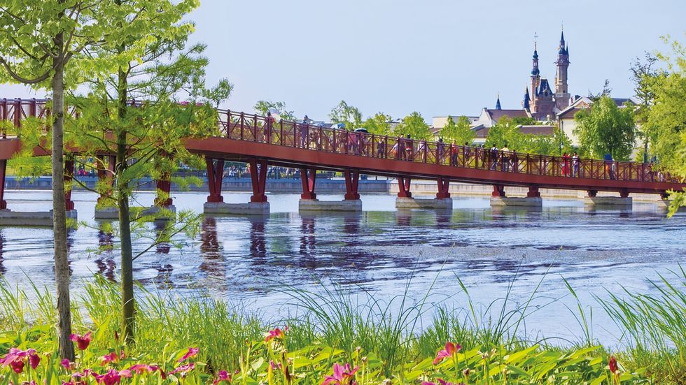Red bridge over a river with green trees and a distant view of building spires at Shanghai Disney Resort