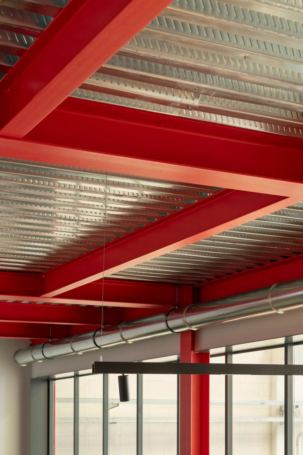 Red metal beams and silver pipes on an industrial ceiling with large windows at Leva Grid.