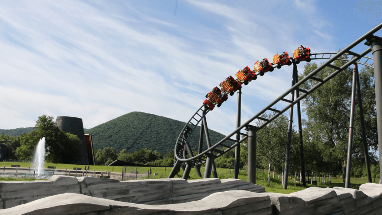 Red roller coaster, Namazu, looping in lush green park, Vulcania, with mountains in background.