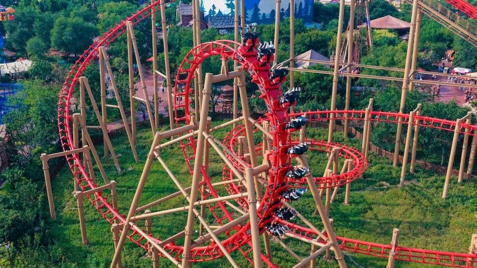 Red roller coaster with loop, surrounded by trees and grass, filled with riders.