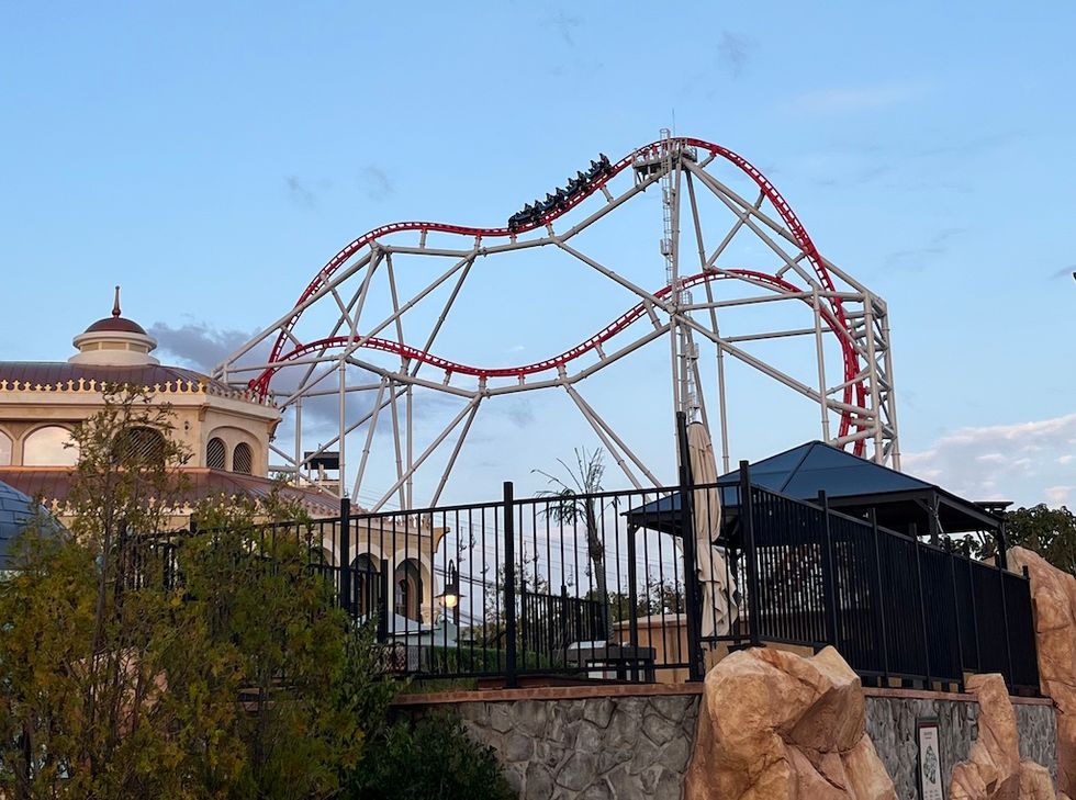 Red roller coaster, Zamperla Double Heart with loops near a building under a blue sky.