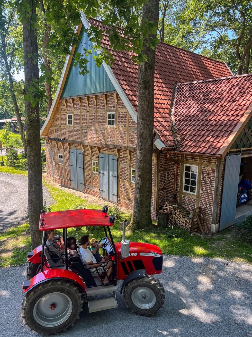 Red tractor at Schloss Dankern, with two passengers in front of a rustic brick barn in a wooded area.