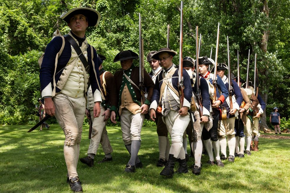Reenactors in colonial military uniforms march on grass with rifles.