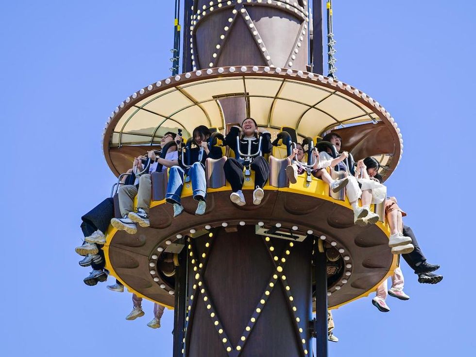 Riders laugh with their legs dangling from a drop-tower ride against a clear blue sky.