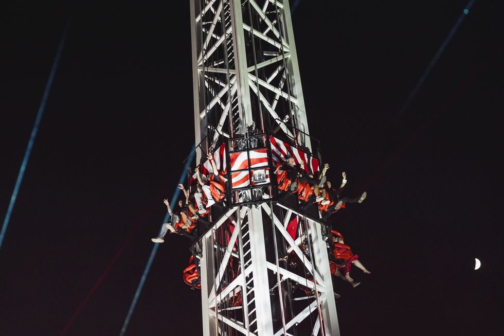 Riders on a drop tower ride at night, with a crescent moon in the background.