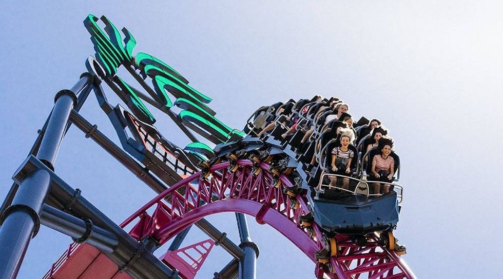 Riders on a steep purple roller coaster drop under a clear blue sky.