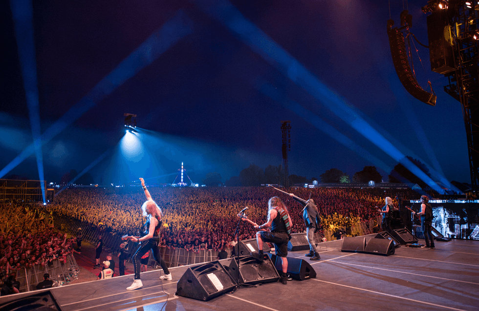 Rock band called Iron Maiden performing live at night with a large, cheering crowd and bright stage lights.