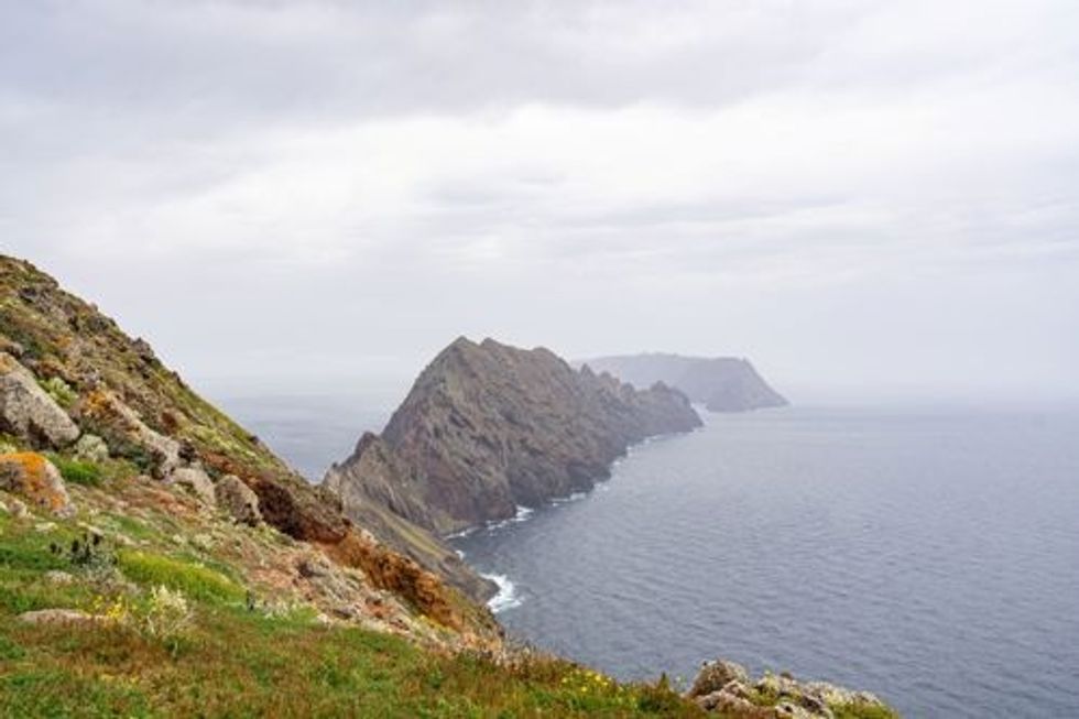 Rocky coastal cliffs overlooking a calm, foggy sea under a cloudy sky.
