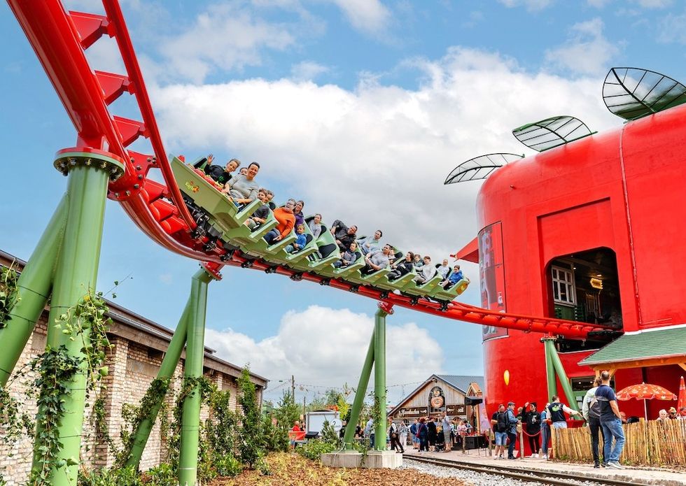 Roller coaster riders pass a giant red building against a partly cloudy sky.