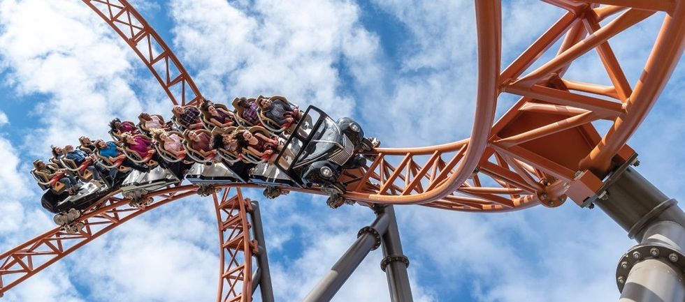 Roller coaster riders thrill on an orange track against a cloudy blue sky.