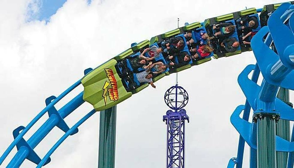 Roller coaster riders twist on a vibrant blue and green track against a cloudy sky.