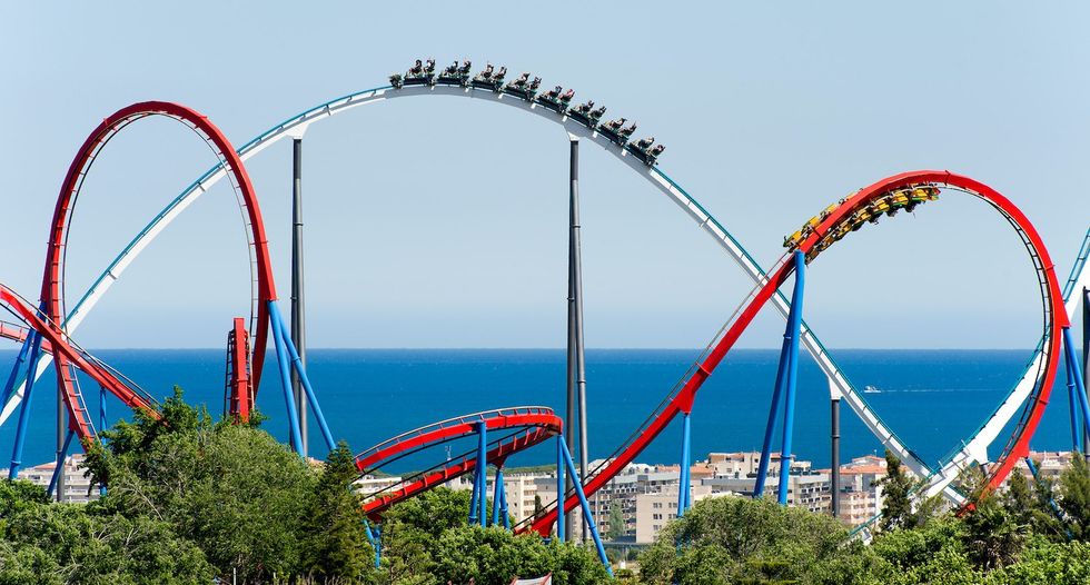 Roller coaster with loops near the ocean, surrounded by trees and city buildings.