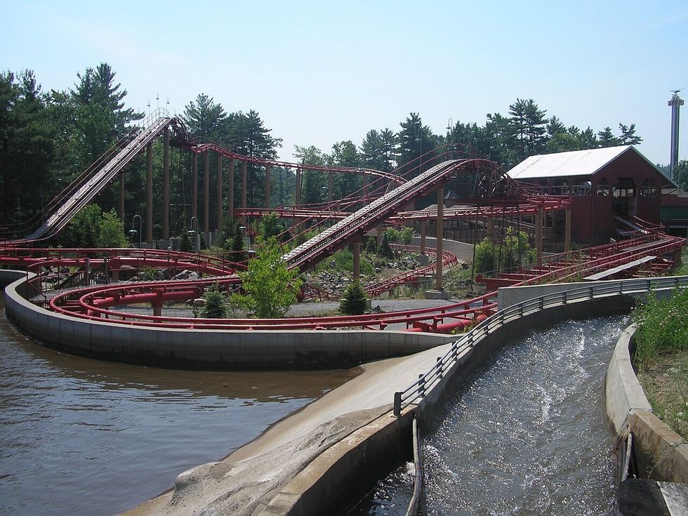 Roller coaster with red tracks and water feature in a wooded amusement park.