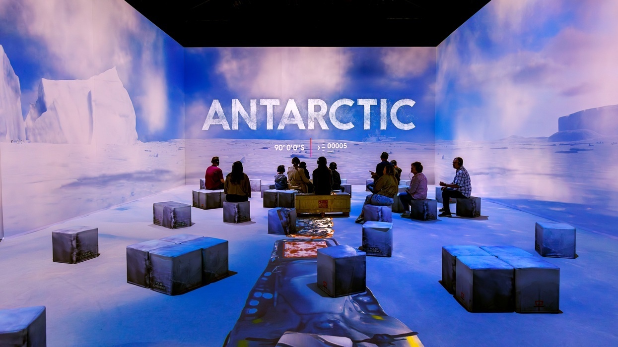 Room with Antarctic projection at Polar Experience, with people seated on cube stools among icy visuals.