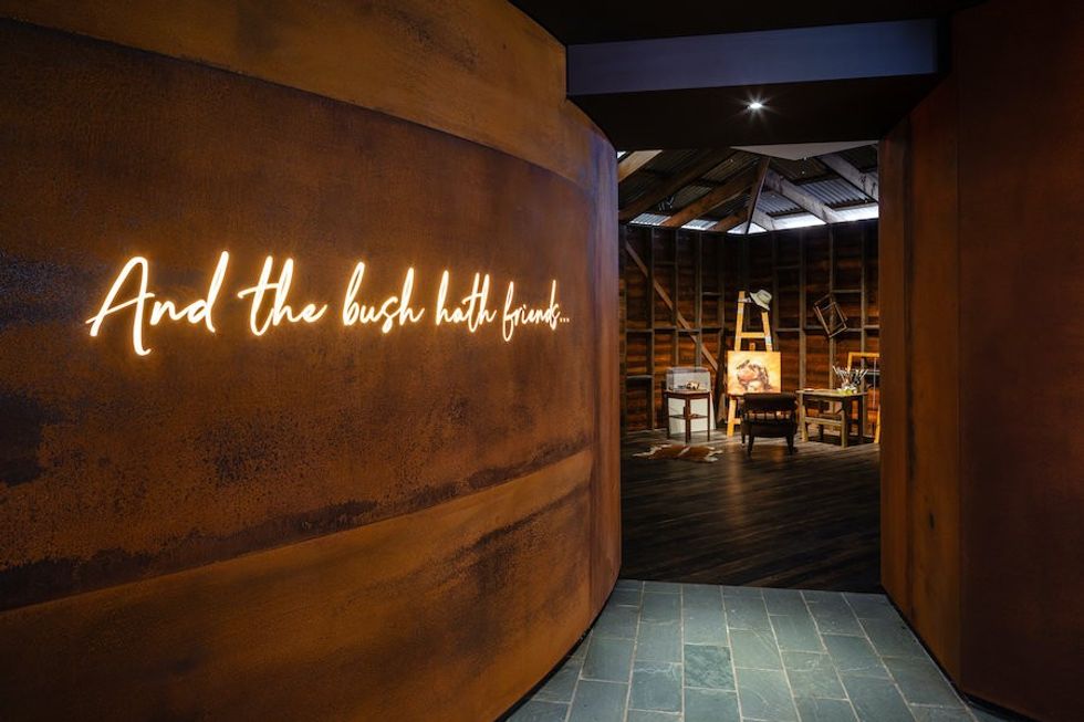 Rust-colored wall leading into gallery with lit text "and the bush hath friends"