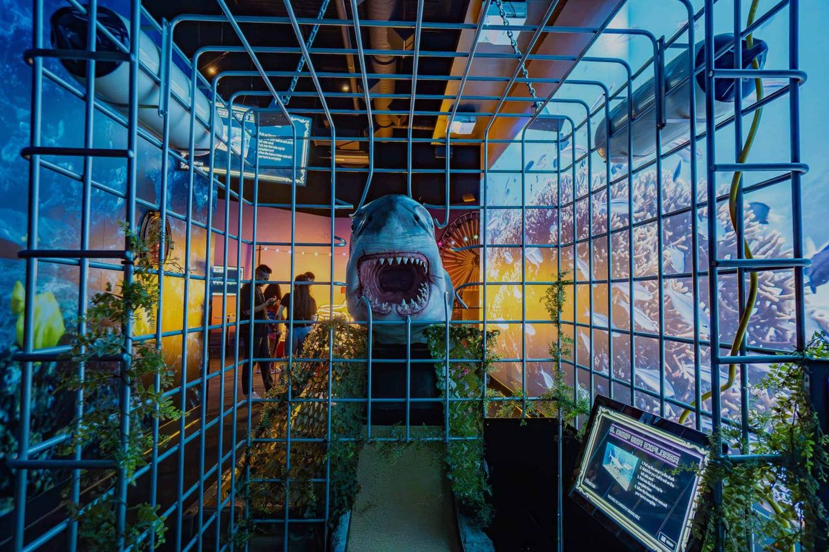 Shark in a cage mini-golf hole with colorful ocean backdrop and visitors observing.