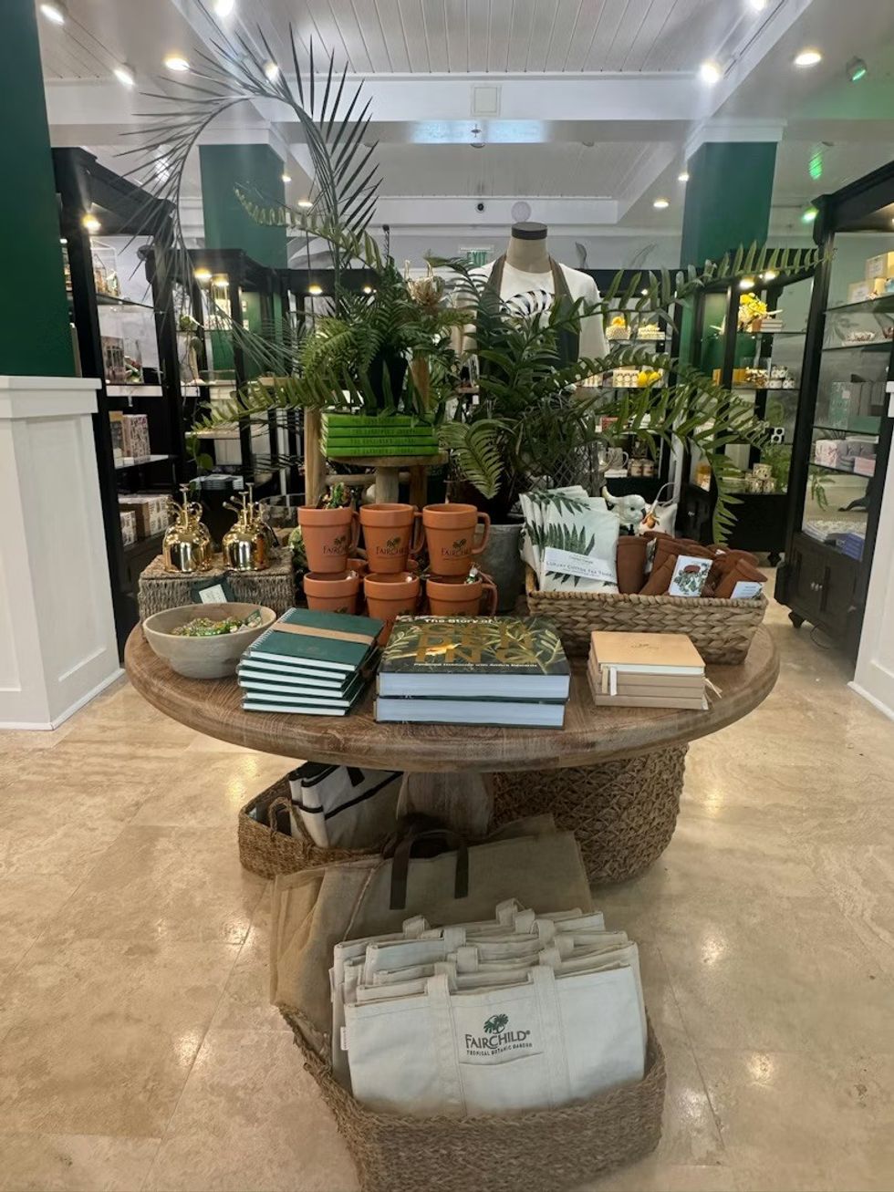 Shop display with plants, books, mugs, and tote bags on a wooden table. The Sanctuary Shop at Fairchild Tropical Botanic Garden