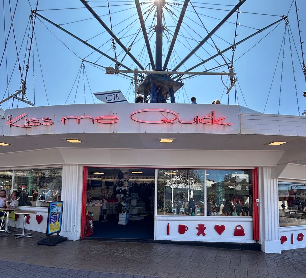 Shop with "Kiss me Quick" in neon, beneath a tall structure with wires above.