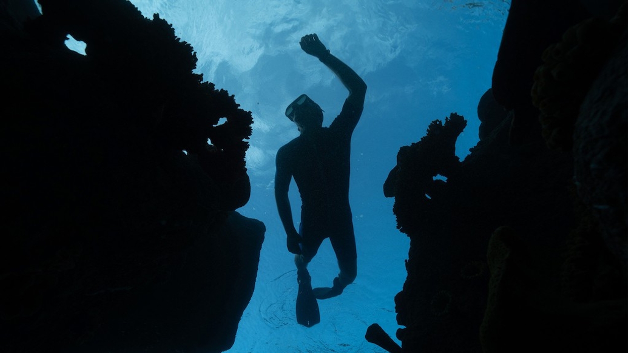 Silhouette of a snorkeler underwater between coral formations, reaching towards the surface.