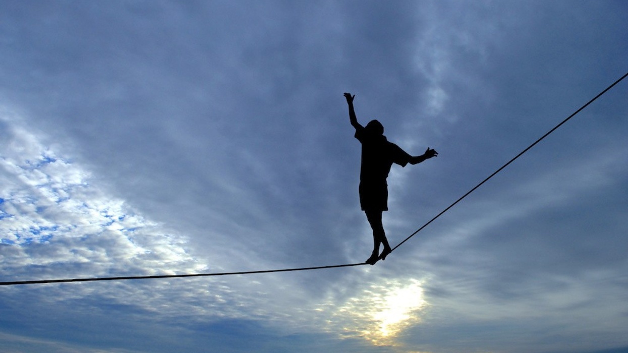 Silhouette of young man balancing on slackline, sun and clouds behind. Slackliner balancing on tightrope between two rocks, highline silhouette.