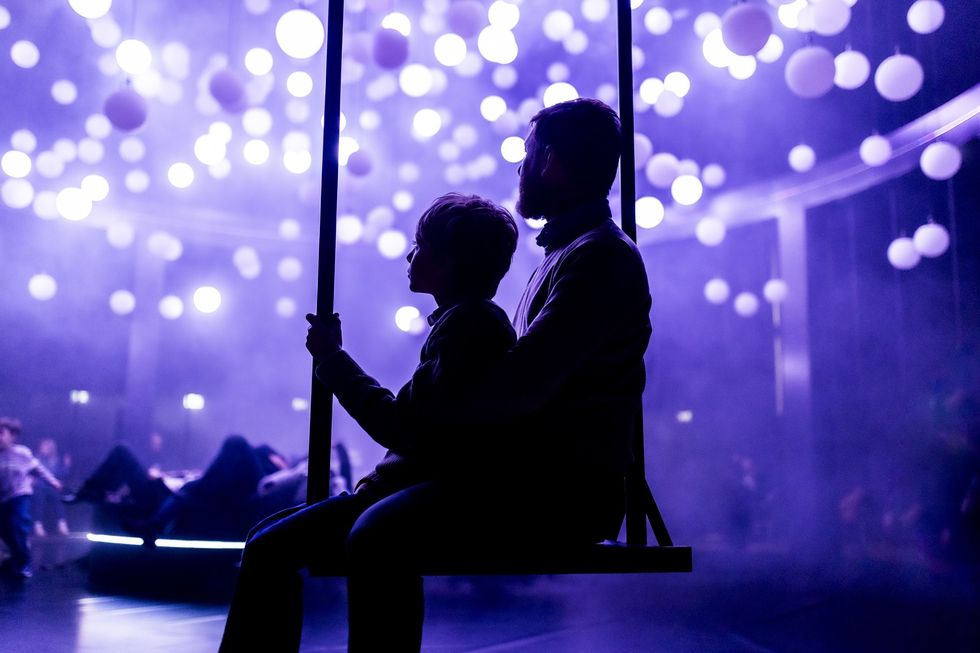 Silhouetted child and adult on swing in a room with purple lighting and hanging spheres.