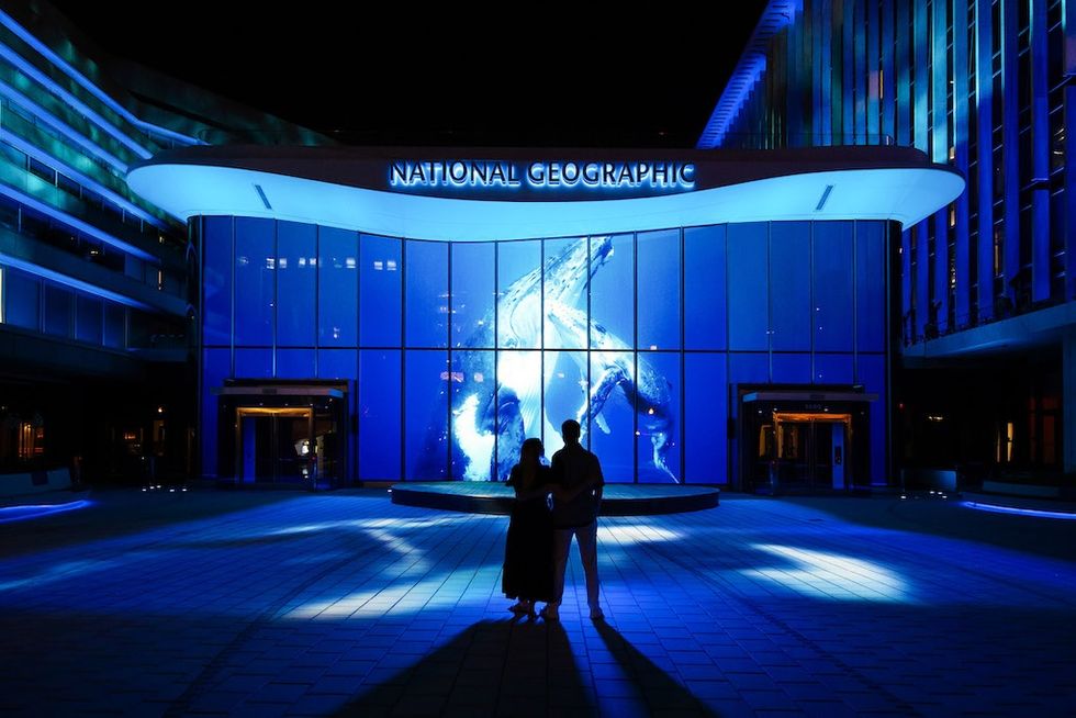 Silhouetted couple in front of lit-up National Geographic building at night.