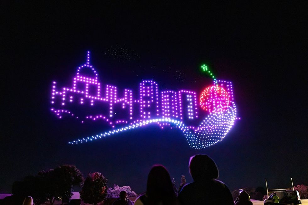 Silhouetted crowd watches colorful drone light display of skyline and cherry.