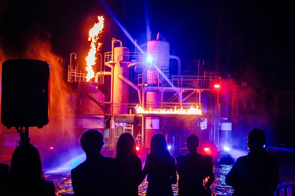 Silhouetted crowd watches fiery industrial performance at night with vibrant lights.