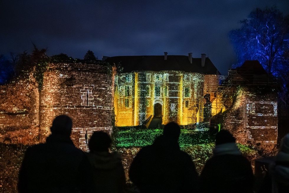 Silhouetted figures watch castle illuminated by colorful lights at night.