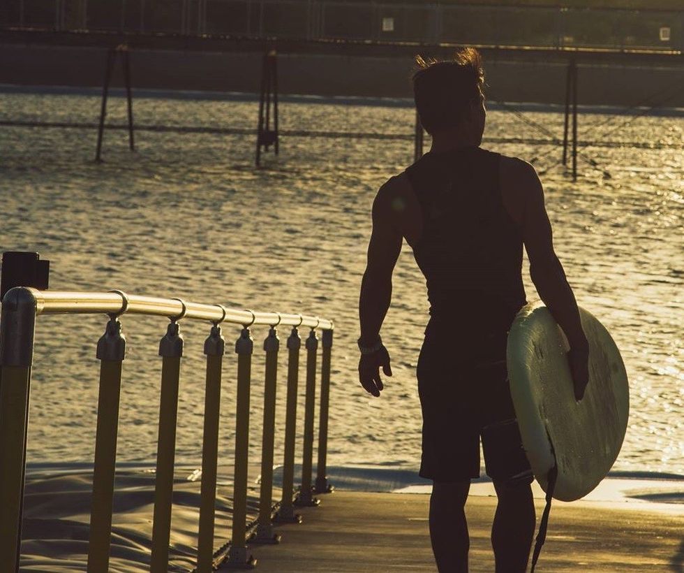 Silhouetted surfer holding a board, walking along a pier at sunset.
