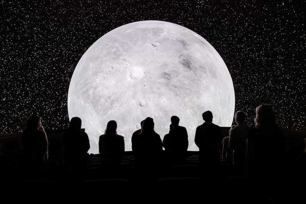 Silhouettes view a large moon projection against a starry background.