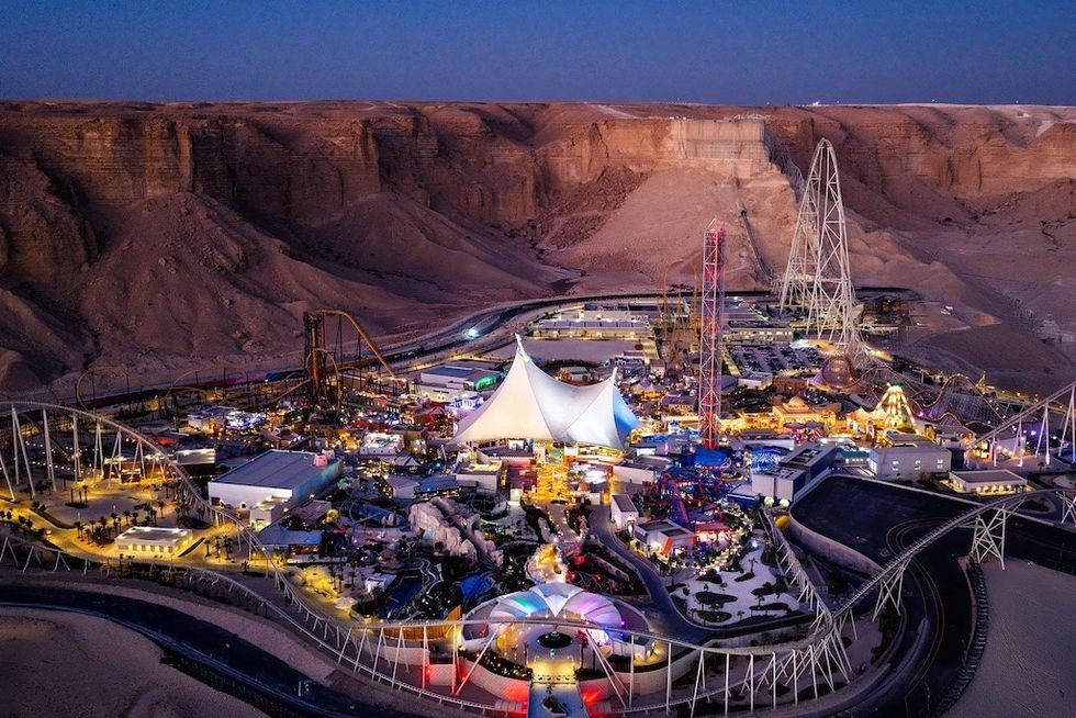 Six Flags Qiddiya at dusk with colorful lights and roller coasters surrounded by rocky desert landscape.