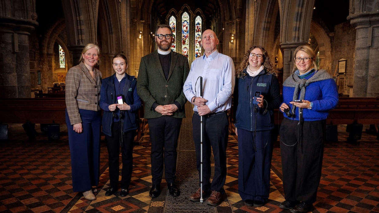 Six people standing in St Canice's Cathedral with stained glass windows in the background, they hold audio tour devices and one person holds a white cane