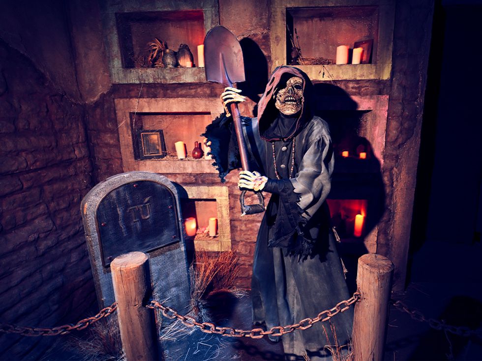 Skeleton in cloak holding shovel in eerie, candle-lit crypt scene.
