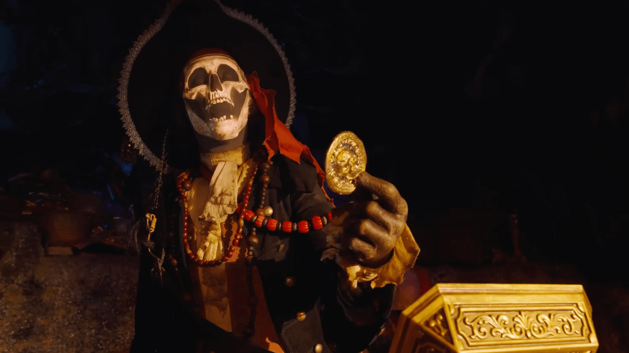 Skeleton pirate at Disneyland holding a gold coin next to an ornate chest in dim light.