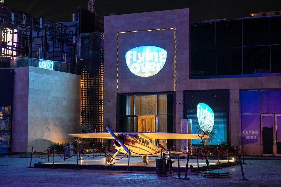 Small airplane displayed outside "Flying Over Saudi" attraction at night.