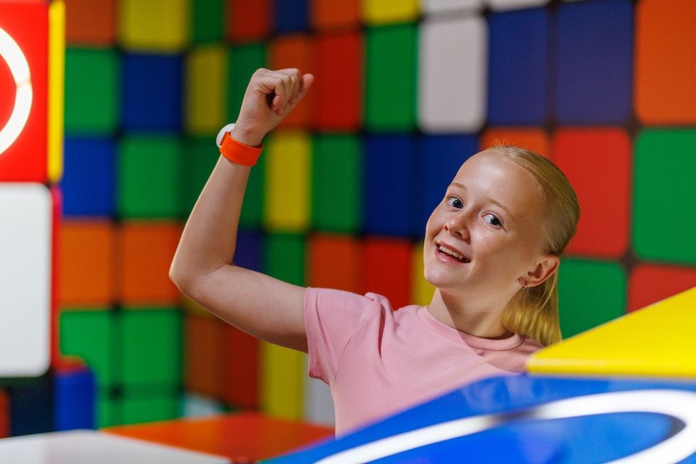 Smiling child in a pink shirt raises arm in colorful, block-patterned play area, wearing Pico Play PlayMAXX RFID band