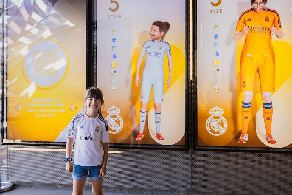 Smiling child in a Real Madrid jersey stands before sports-themed digital displays.