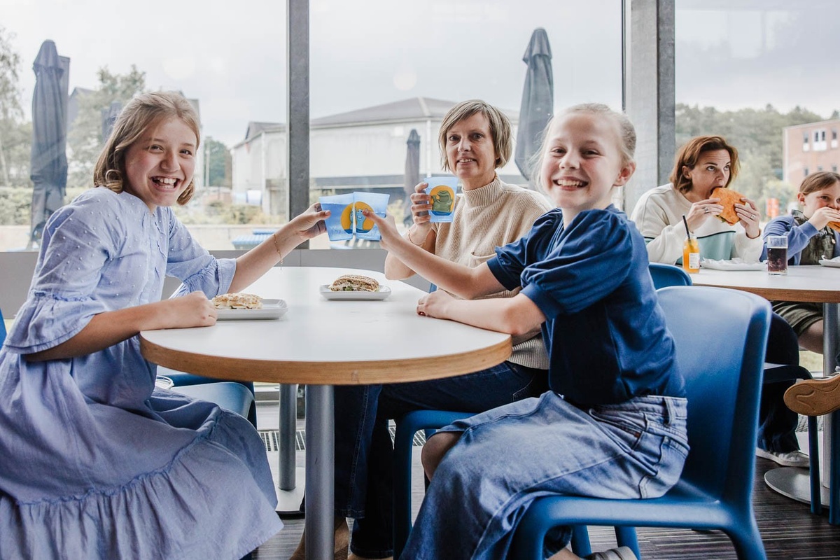 Smiling children in a cafe sharing a cheerful moment over a meal with adults in the background.