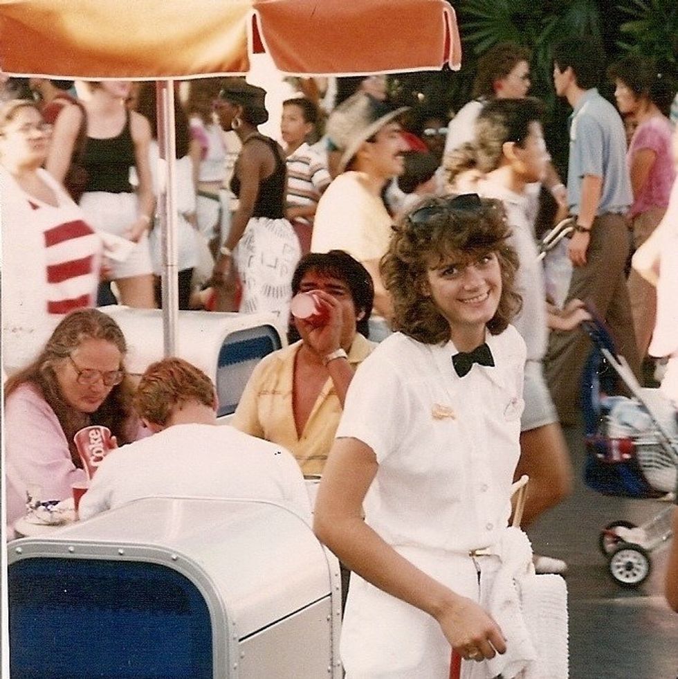 Smiling employee, Cheryl Christensen as a teen, in a white uniform with a bow tie among a busy crowd at Disneyland