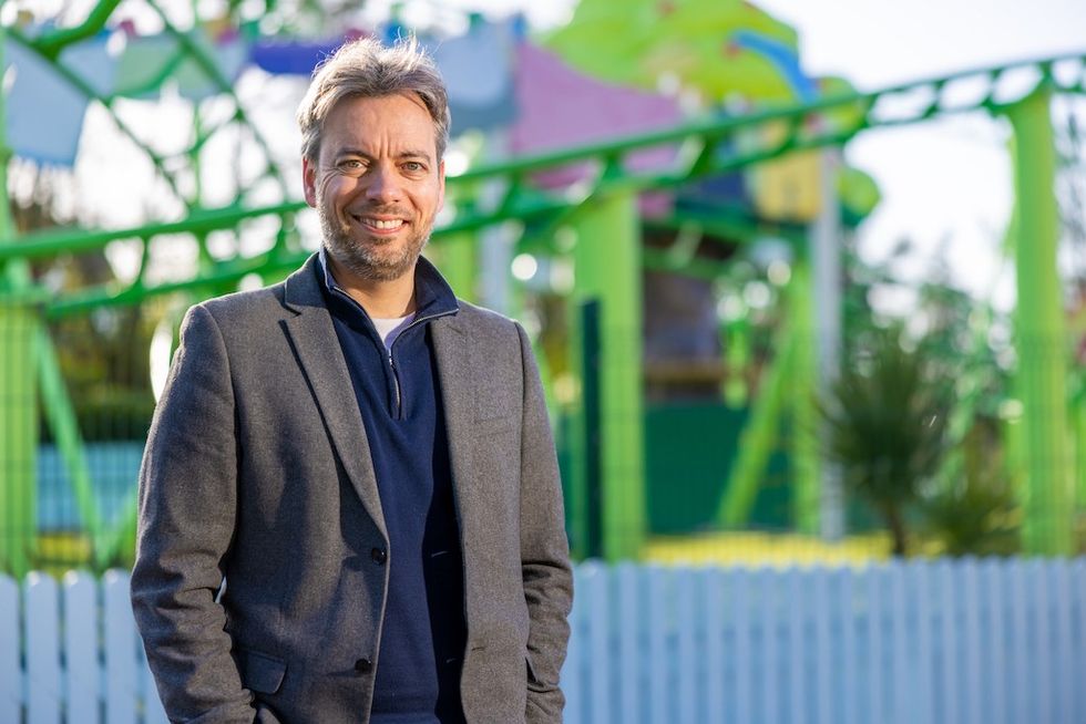Smiling man in a blazer stands outdoors with colorful amusement park ride behind.