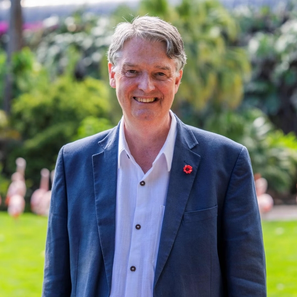 Smiling man in a blazer with flamingos and greenery in the background.