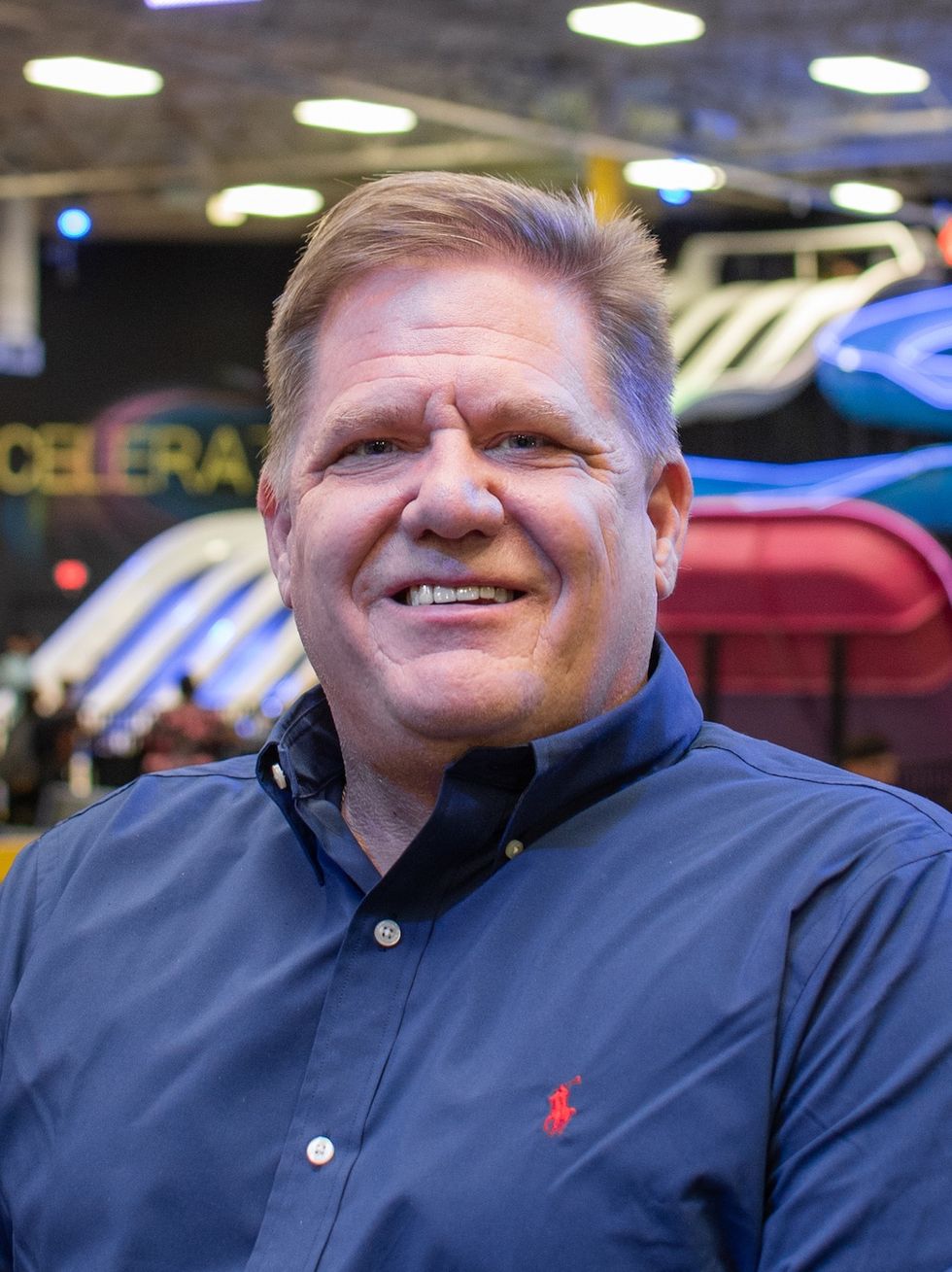 Smiling man in a blue shirt at an indoor venue with colorful lights in the background.