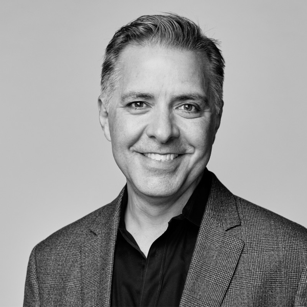 Smiling man in a checked blazer and dark shirt against a plain backdrop.